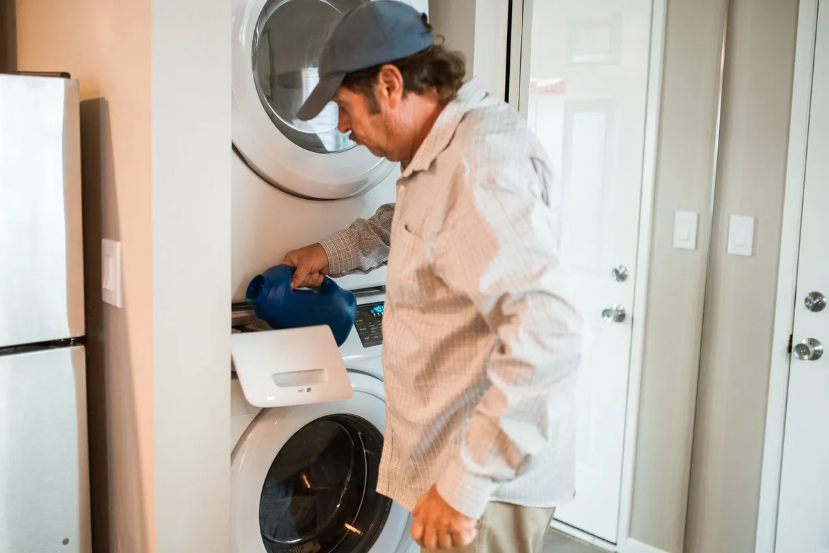 Man pouring detergent into a stacked washer and dryer in a modern laundry room, emphasizing appliance care.